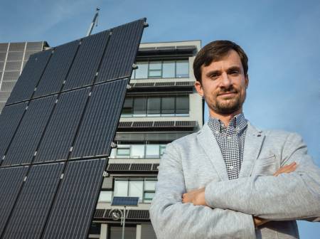 Ein bärtiger junger Mann steht mit verschränkten Armen neben einer PV Anlage und schaut entschlossenen Blickes in die Kamera. 