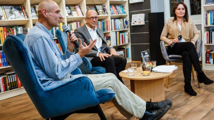 Zwei Männer und eine Frau auf einem Podium in Stühlen sitzend, im Hintergrund eine Bücherwand