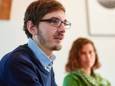 Ein Mann mit Brille schaut nach links, im Hintergrund ist unscharf die Frau mit dem grünen Pullover zu sehen.