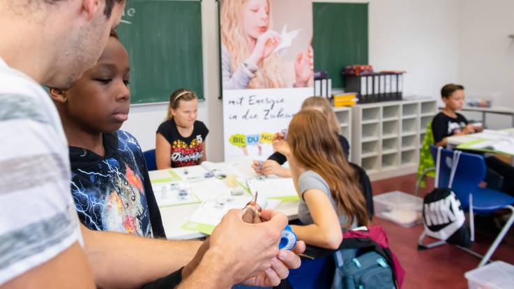 Ein  junger Mann erklärt einem Jungen etwas. Im Hintergrund der Schulraum, Kinder am Tisch.