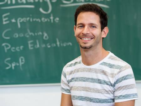 Halbportrait eines jungen Mannes vor einer Schultafel.