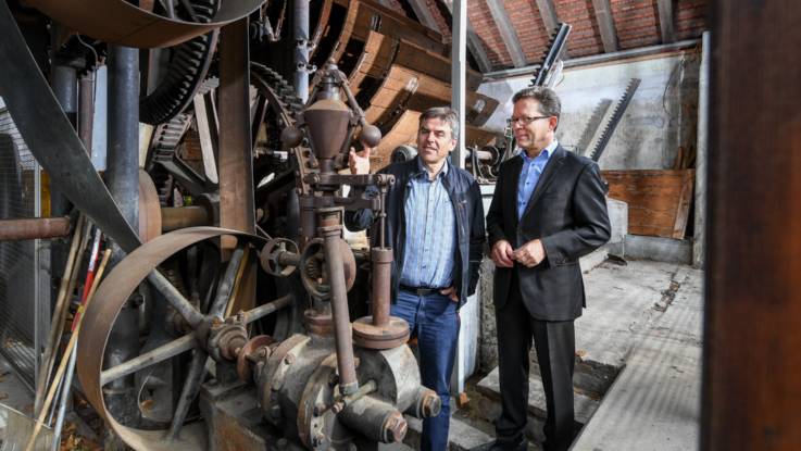 Zwei Männer stehen in der Anlage der historischen Wasserkraftanlage mit großen Zahnrädern, die Wasserschaufelräder im Hintergrund