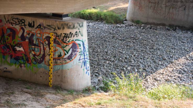 Ein Pegelstandsanzeiger am Betonträger einer Brücke, die über ein ausgetrocknetes Flussbett mit großen Kieseln führt.