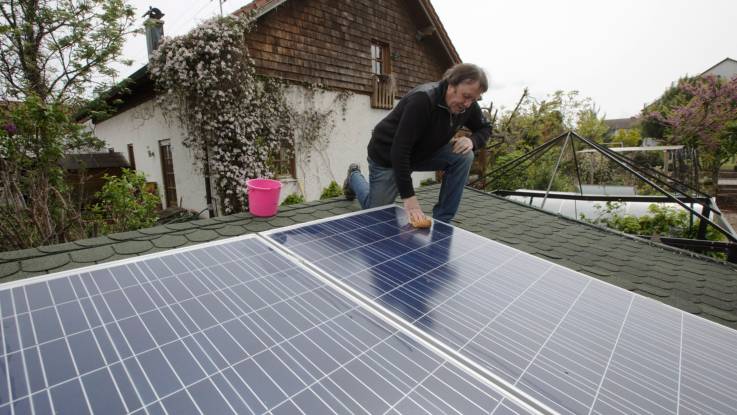 Auf dem Dach eines Gartenhäuschens putzt ein Mann mit Wassereimer und Schwamm ein PV-Modul.