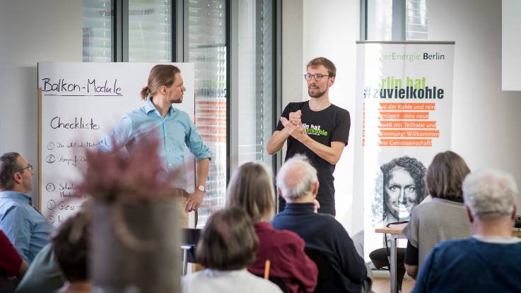 Blick auf zwei Vortragende vom Publikum aus; im Hintergrund sind Stelltafen mit den Überschriften «Balkon-Module» und «Berlin hat zu viel Kohle» zu erkennen. 