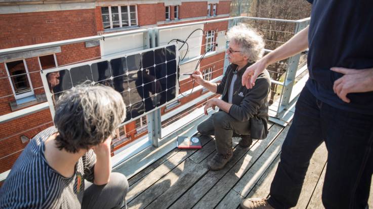 Balkonszene: In der Bildmitte hantiert ein Mann mit strubbeligen grauen Haaren an einem PV-Modul, zwei weitere Menschen schauen dabei zu.