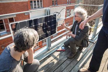 Balkonszene: In der Bildmitte hantiert ein Mann mit strubbeligen grauen Haaren an einem PV-Modul, zwei weitere Menschen schauen dabei zu.