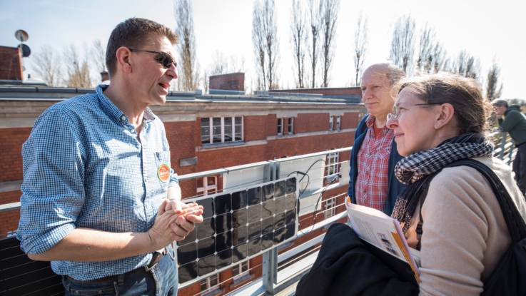 Vor einem Solarmodul an einem Balkongeländer diskutiert ein Mann mit Kurzhaarschnitt und Sonnenbrille mit zwei Interessenten.