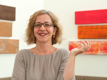 Symphatische junge Frau mit blonder Mähne und Brille, vor einem Kunstwerk fotografiert.