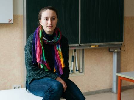 Eine junge Frau sitzt in einem Klassenraum auf einem Tisch und schaut in die Kamera.