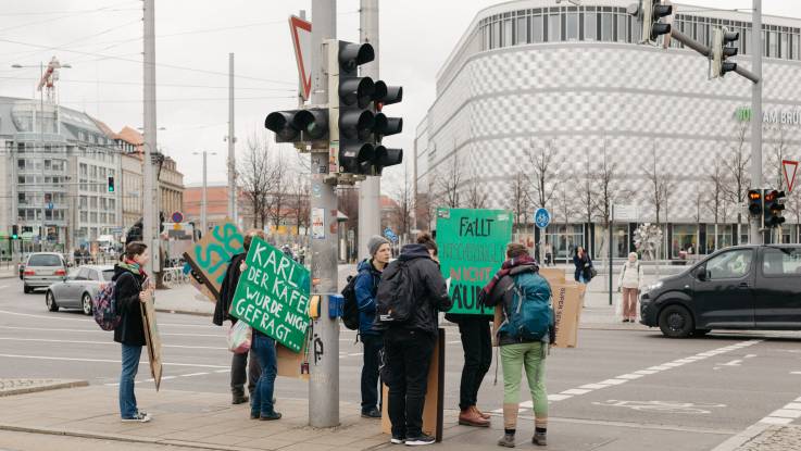 Ein paar junge Leute mit Demoplakaten an einer Straßenkreuzung, im Hintergrund die denkmalgeschützte Aluminium-Fassade des ehemaligen Konsument-Warenhauses am Brühl, Leipzig.