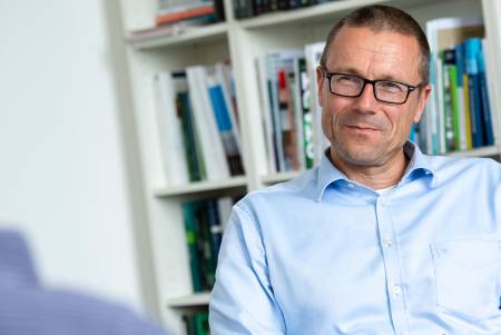 Ein lächelnder Mann sitzt entspannt am Tisch, die Hände locker im Schoß liegend.