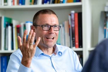 Ein Mann mit Brille und aufgeknöpftem Hemd hebt im Gespräch gestikulierend die rechte Hand.