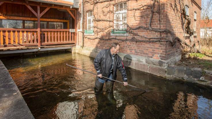 Ein Mann mit Gummistiefeln steht mit einer Hakenstange in einem kleinen Wasserkanal.