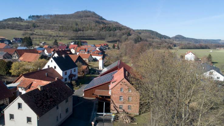 Luftbild eines großen Backsteingebäudes mit zwei Anbauten, deren Dächer auf einer Seite vollflächig mit PV-Modulen belegt sind.