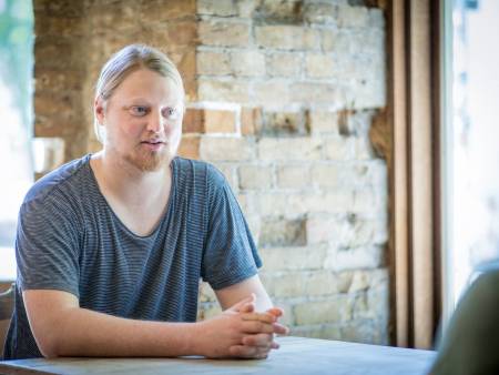 Ein junger Mann mit blonden Haaren und Zopf sitzt an einem Tisch und spricht.