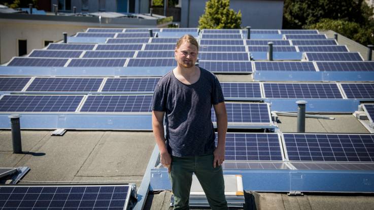 Ein junger Mann mit blondem Zopf steht zwischen PV-Paneelen auf einem Flachdach. 