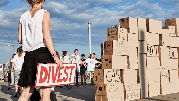 Berlin im Sommer: Eine Pappkartonmauer. Auf den Kartons steht «Öl», «Gas» und «Kohle» geschrieben. Davor eine Frau, die ein Schild mit der Aufschrift «Divest» in der Hand hält.