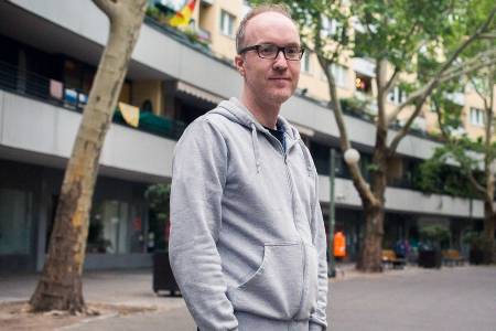 Ein junger Mann mit Brille und grauem Hoody steht auf einem städtischen Platz.