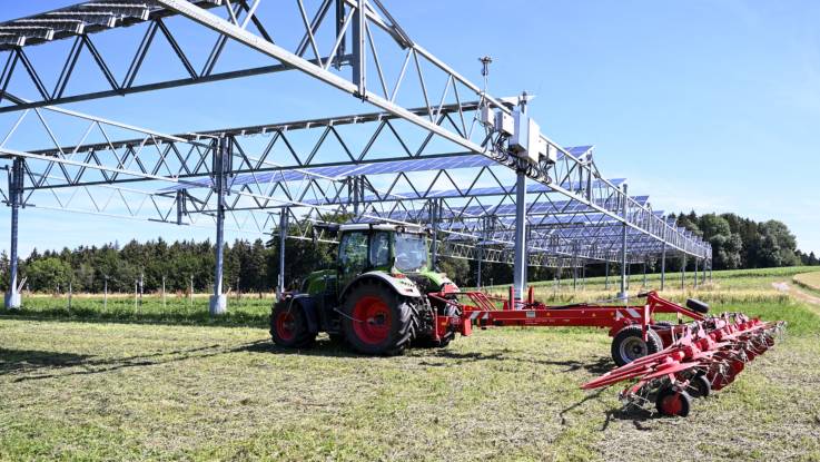 Auf einem Acker steht ein Tragwerk, auf dem PV-Module montiert sind, darunter fährt ein Traktor mit einem Heuwender.