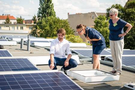 Auf dem Flachdach hockend, zeigt und erläutert ein junger Mann zwei daneben stehenden Frauen ein PV-Modul.