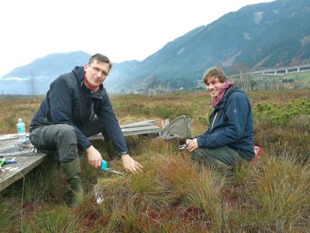 Zwei Männer sitzen in einer flach bewachsenen Landschaft – im Hintergrund Berge – und schneiden Wollgraspflanzen ab.