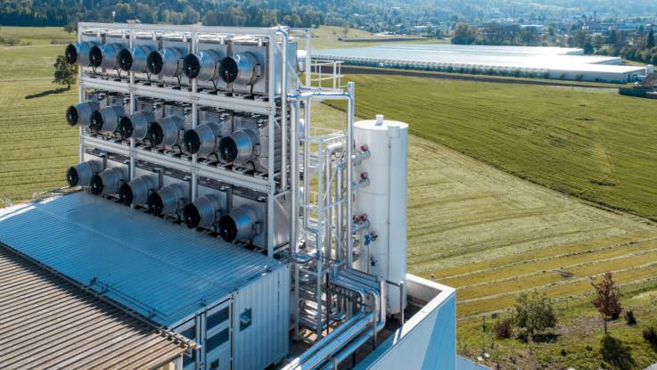 Auf einem alleinstehenden Gebäude im Grünen sind auf einem Tragegestell 18 große Ventilatoren in drei Reihen installert, Rohrleitungen führen von den Ventilatoren zu einem weißen, zylindrischen Gastank.