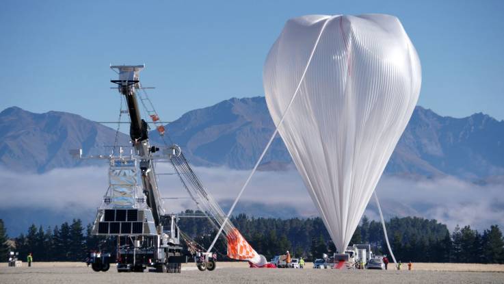 Vor einer Bergkulisse wird ein silbriger Heißluftballon von der Größe eines zehnstöckigen Hauses aufgerichtet, daneben steht ein großer hydraulischer Kran, an dem PV-Module angebracht sind. 