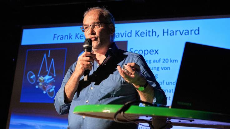 Ein mittelalter Mann mit Mikrofon in der Hand referiert auf der Bühne.