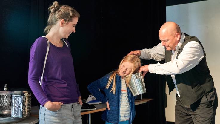 Ein smyphatischer Mann im Hemd und Weste scherzt mit einen Mädchen, dem er einen Champagnerkühler an das Ohr hält. Die Mutter des Mädchen schaut amüsiert zu.