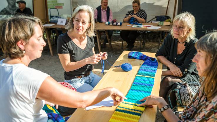 Draußen sitzen vier Frauen um einen Tisch herum, auf dem ein bunt gestreifter Schal liegt, an dem eine der Frauen strickt. Die Frauen sind im Gespräch miteinander.