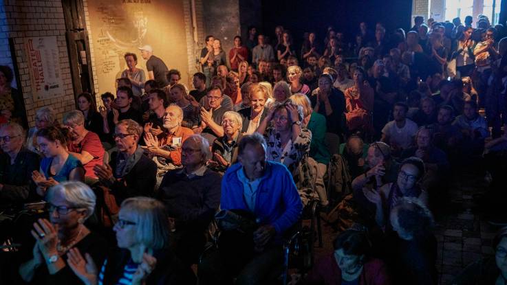 Ein gemischtes Publikum im Halbdunkel im voll besetzen Saal.