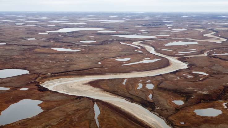 Luftbild einer rostbraunen, kargen Landschaft mit vielen kleinen Wasserflächen und einem Fluss.