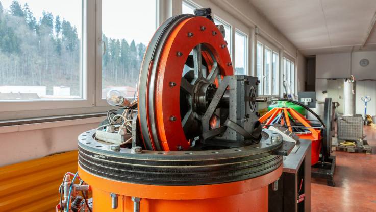 Eine Reihe orange farbener Maschinen in einer kleinen Fabrikationshalle, mit Blick auf einen winterlichen Wald.