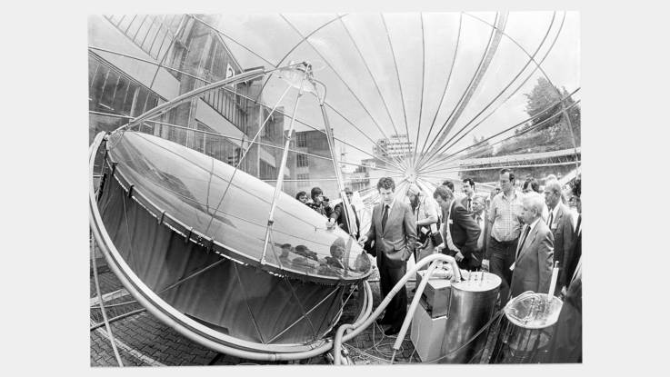 Neben einem ca 3 Meter im Durchmesser großen Parabolspiegel stehen interessierte Männer.