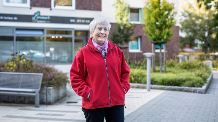 Eine sympathische ältere Dame steht auf einem Platz, die Hände in den Taschen ihrer roten Fleecejacke.