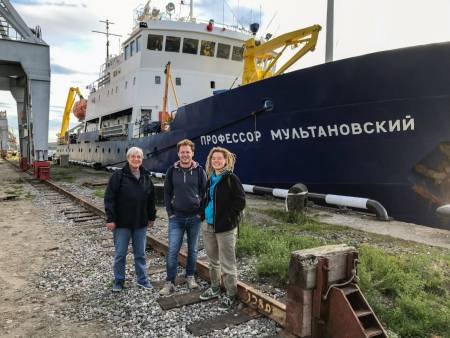 Ein großes blau-weißes Schiff liegt im Hafen, davor posieren drei Personen.