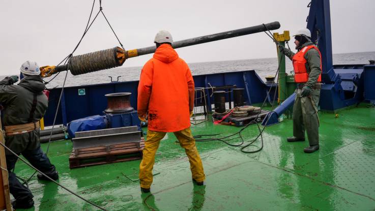 Zwei Männer in Schutzkleidung auf dem Schiffsdeck manövrieren ein an einem Seil hängendes langes Lot, das eine Verdickung in der Mitte hat.