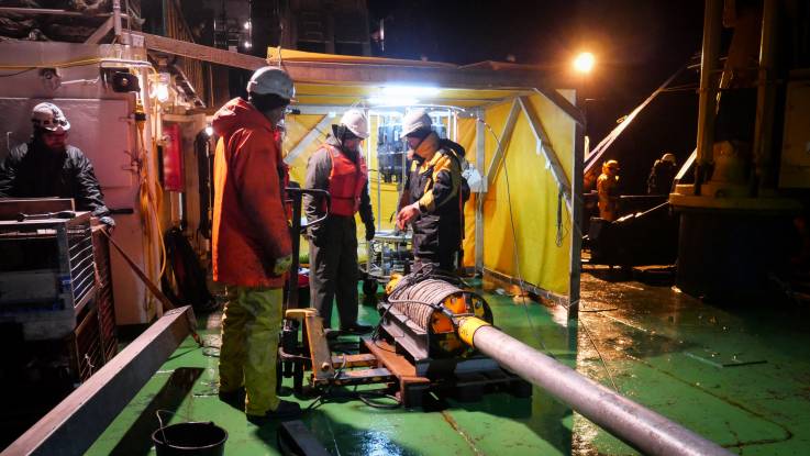 In der Nacht auf dem Schiffsdeck, beleuchtet von Scheinwerfern, arbeiten mehrere Männer an einem liegenden Schwerelot.