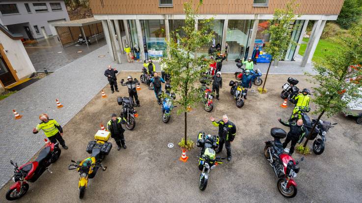 Foto aus der Vogelperspektive: In einem Innenhof steht eine Gruppe Biker neben ihren Maschinen und winkt nach oben in die Kamera.