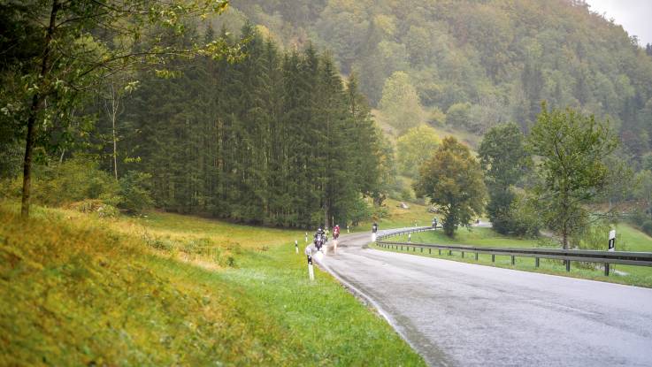 Auf einer idyllischen Landstraße in den Bergen im strömenden Regen fährt eine kleine Motorradkolonne.