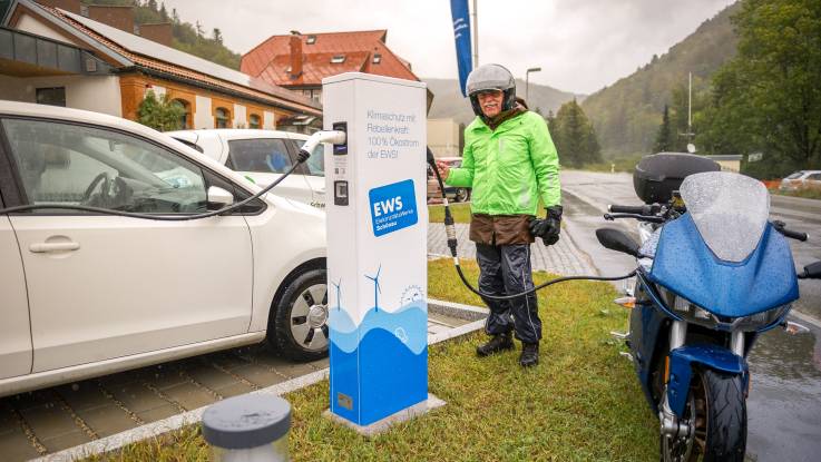 Im Regen steht ein Mann in Motorrradkluft an einer Ladesäule mit Aufdruck der EWS und lädt sein Mototrrad.