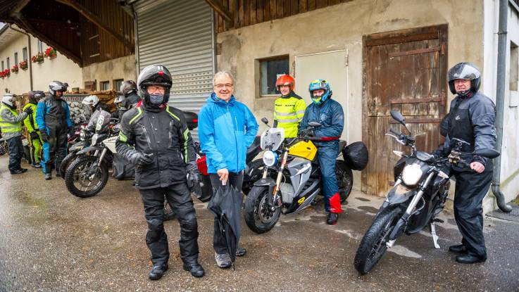 Unter einem historischen Hausdach steht eine Gruppe Motorradfahrer und ein Mann mit Regenschirm.