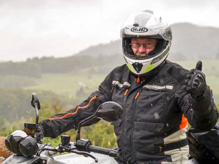 Im Regen sitzt ein Mann in Regenkleidung auf einem großen BMW-Motorrad, lacht in die Kamera und hebt den Daumen.