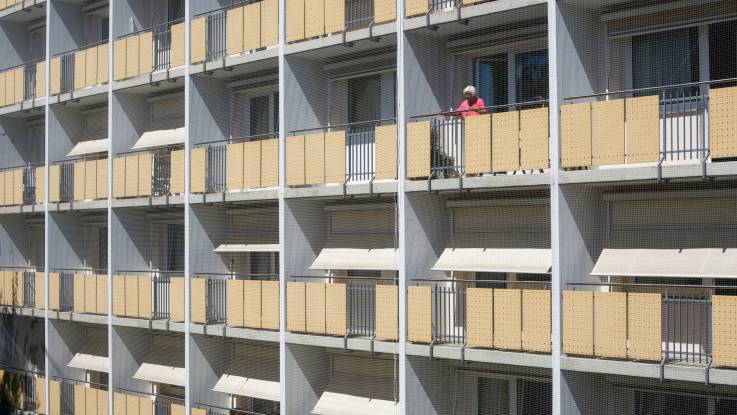 Die Sonne scheint auf eine Hausfassade mit Balkonen, auf einem der Balkons steht eine alte Dame.