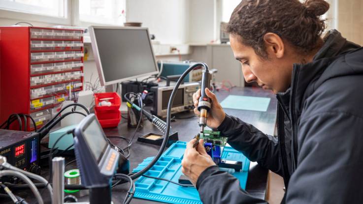 Ein junger Mann sitzt an einem Werktisch mit elektronischen Geräten und lötet an einer Platine