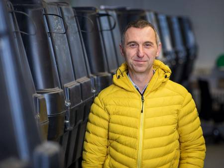 Ein älterer Mann mit kurzen Haaren und gelber Daunenjacke steht in einer Halle neben schwarzen Plastikbehältern.