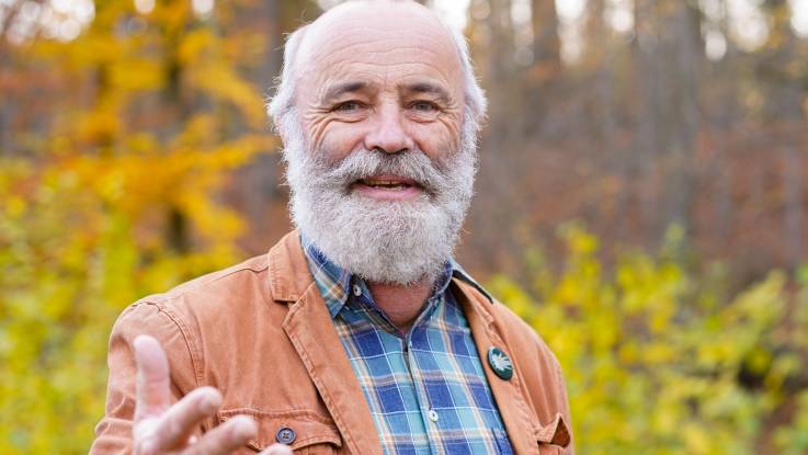 In einem herbstlich bunten Wald steht ein älterer grauhaariger Mann mit einem Vollbart, gestilkuliert und schaut in die Kamera.