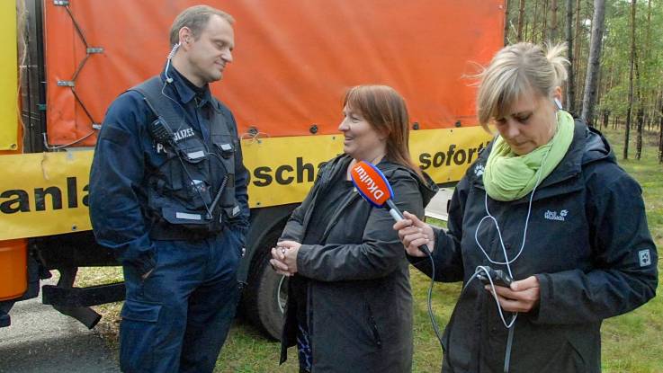 Vor einem LKW steht ein Polizist und spricht mit einer Aktivistin. Einer junge Frau rechts daneben hält ein Mikrofon mit Aufdruck «DRadio» in ihre Richtung und nimmt auf. 