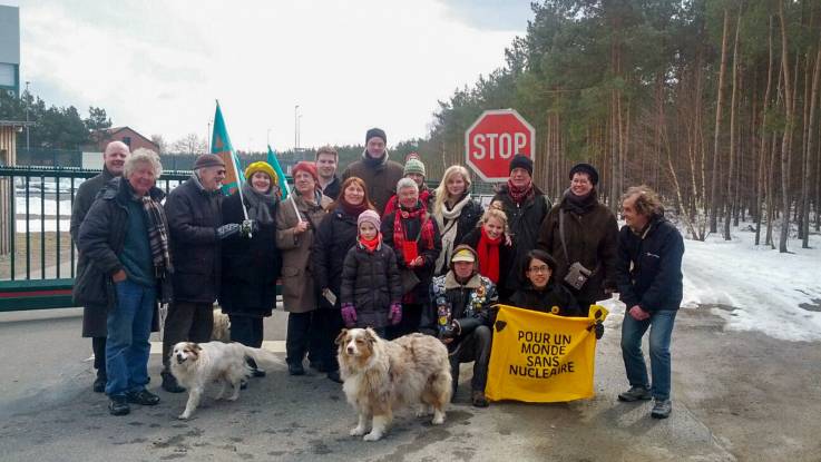 Vor einem gut abgesperrten Gelände steht eine große Gruppe winterlich gekleideter Menschen mit zwei Hunden und einem Transparent, auf dem steht: «Pour un monde sans nucleaire»."